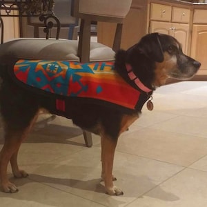 May include: A black and brown dog wearing a colourful patterned fleece vest with a pink collar and a tag. The dog is standing on a tiled floor.