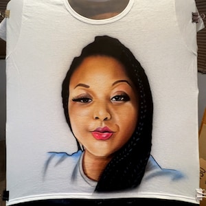 K&ouml;nnte beinhalten: Wei&szlig;es T-Shirt mit einem detaillierten Portr&auml;t einer Frau mit dunkler Haut, Z&ouml;pfen und rotem Lippenstift. Das Kunstwerk ist auf dem T-Shirt zentriert und zeigt das Gesicht und die Schultern. Das T-Shirt wird mit Clips aufgeh&auml;ngt.