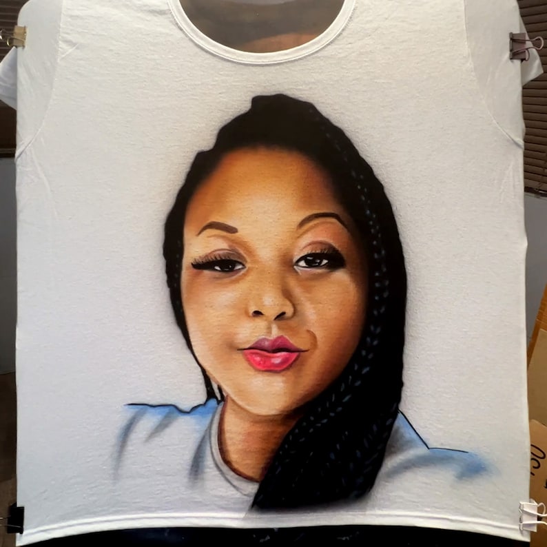 K&ouml;nnte beinhalten: Wei&szlig;es T-Shirt mit einem detaillierten Portr&auml;t einer Frau mit dunkler Haut, Z&ouml;pfen und rotem Lippenstift. Das Kunstwerk ist auf dem T-Shirt zentriert und zeigt das Gesicht und die Schultern. Das T-Shirt wird mit Clips aufgeh&auml;ngt.