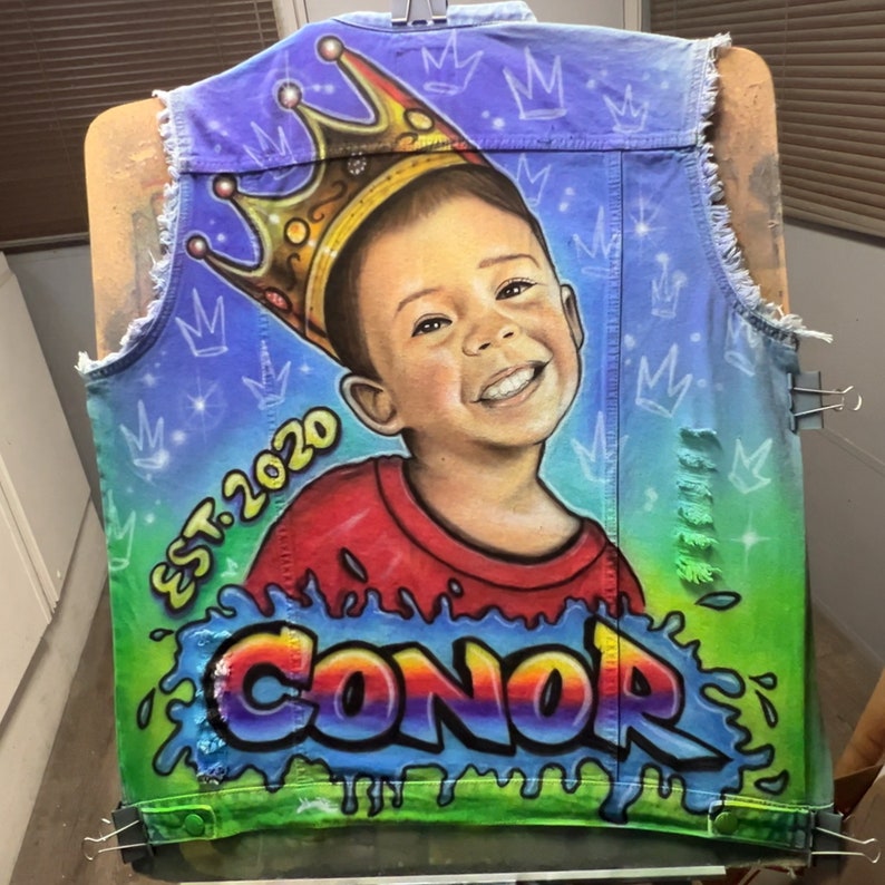 K&ouml;nnte beinhalten: Eine Jeansweste mit einem individuellen Airbrush-Portr&auml;t eines l&auml;chelnden Kindes mit Krone. Die Weste tr&auml;gt den Namen "CONOR" im Graffiti-Stil, zusammen mit "EST. 2020" und Kronengrafiken. Der Hintergrund ist ein Farbverlauf aus Blau, Gr&uuml;n und Lila.