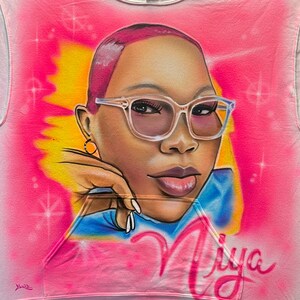 K&ouml;nnte beinhalten: Rosa Kapuzenpullover mit einem Airbrush-Portr&auml;t einer Person mit Brille. Das Portr&auml;t ist vor einem rosa und gelben Hintergrund mit dem Namen "Niya" in Schreibschrift platziert. Der Hoodie hat eine Vordertasche.