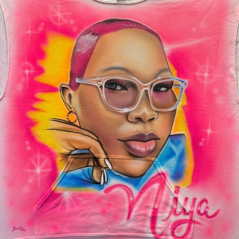 K&ouml;nnte beinhalten: Rosa Kapuzenpullover mit einem Airbrush-Portr&auml;t einer Person mit Brille. Das Portr&auml;t ist vor einem rosa und gelben Hintergrund mit dem Namen "Niya" in Schreibschrift platziert. Der Hoodie hat eine Vordertasche.