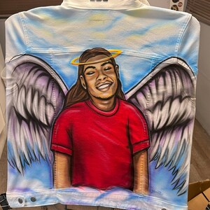 K&ouml;nnte beinhalten: Wei&szlig;e Jeansjacke mit einem Airbrush-Design, das eine l&auml;chelnde Person mit Heiligenschein, Engelsfl&uuml;geln und einem roten Hemd zeigt. Der Hintergrund ist ein blauer Himmel mit Wolken. Die Jacke hat einen Knopfverschluss.