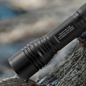 May include: Black flashlight with a textured grip and a smooth, cylindrical body. The flashlight has the text "THANK YOU FOR LIGHTING OUR PATH TO ARROW OF LIGHT!" on the side. The flashlight is resting on a piece of wood.