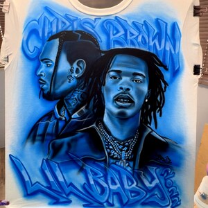 K&ouml;nnte beinhalten: Wei&szlig;es T-Shirt mit Airbrush-Portr&auml;ts von Chris Brown und Lil Baby in Blaut&ouml;nen. Die W&ouml;rter "CHRIS BROWN" und "LIL BABY" sind ebenfalls im Graffiti-Stil in Blau aufgespr&uuml;ht.
