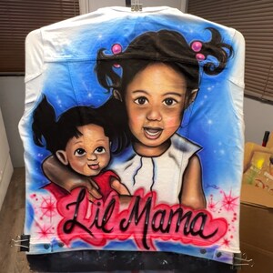 K&ouml;nnte beinhalten: Wei&szlig;e Jeansjacke mit einem Airbrush-Portr&auml;t von zwei Kindern vor blauem Himmel. Die Worte "Lil Mama" sind in roter und schwarzer Schrift am unteren Rand der Jacke geschrieben.