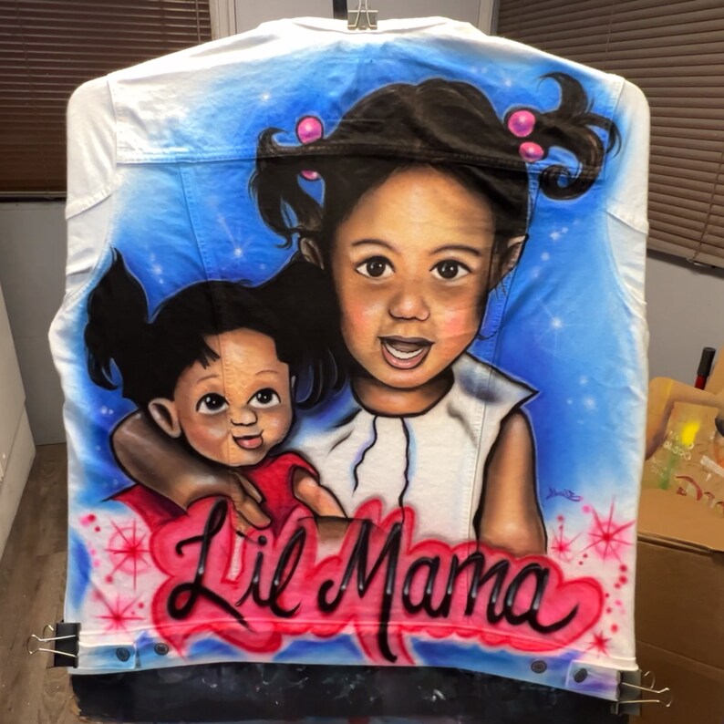 K&ouml;nnte beinhalten: Wei&szlig;e Jeansjacke mit einem Airbrush-Portr&auml;t von zwei Kindern vor blauem Himmel. Die Worte "Lil Mama" sind in roter und schwarzer Schrift am unteren Rand der Jacke geschrieben.