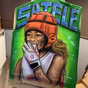 K&ouml;nnte beinhalten: Eine gr&uuml;ne Weste mit dem Wort "SATELE" in blauen Buchstaben. Die Weste zeigt ein gemaltes Portr&auml;t einer Person, die einen orangefarbenen Helm und wei&szlig;e Handschuhe tr&auml;gt. Der Hintergrund ist gr&uuml;n mit wei&szlig;en Glitzer.