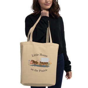 Puede incluir: Bolso tote beige con el texto "Little House on the Prairie" y una ilustraci&oacute;n de una carreta cubierta y caballos. El bolso lo sostiene una persona que lleva una camisa negra de manga larga y vaqueros azules.