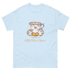 Puede incluir: Camiseta azul claro con una ilustración de acuarela de una taza de té y un platillo. La taza y el platillo tienen rosas rosas y detalles dorados. El texto "My Tea Shirt" está escrito en cursiva. Cuello redondo.