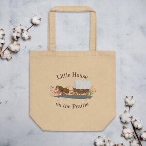 Puede incluir: Bolso tote beige con el texto "Little House on the Prairie" y una ilustraci&oacute;n de una carreta cubierta tirada por dos caballos. El bolso tiene dos asas y est&aacute; hecho de un material similar a la lona.
