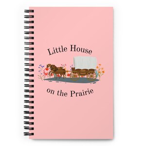 Puede incluir: Un cuaderno de espiral rosa con el texto "Little House on the Prairie". La ilustración de la portada muestra una carreta cubierta tirada por dos caballos marrones, con flores coloridas.