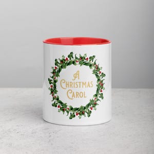 Puede incluir: Taza de cerámica blanca con interior rojo. La taza presenta un diseño de corona verde con bayas rojas y el texto "A Christmas Carol" en letras doradas. Una taza con temática festiva.
