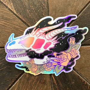 May include: A holographic sticker featuring a skull with a galaxy background and roses. The skull is white with pink and orange accents. The roses are orange and pink with green leaves. The sticker is surrounded by a black border. The text "Amanda Tulacz" is visible at the bottom of the sticker.