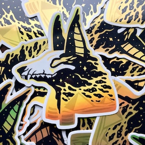 Pu&ograve; includere: Un adesivo nero e oro con un'immagine stilizzata di una testa di sciacallo Anubis su uno sfondo di cielo stellato e piramidi in primo piano.
