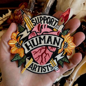 May include: Embroidered patch with a heart, pencils, paintbrush, and flowers. The text reads "Support Human Artists".