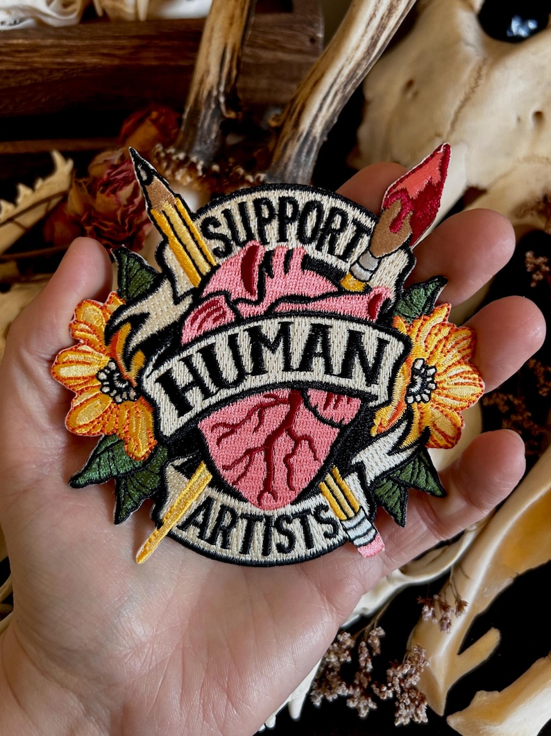 May include: Embroidered patch with a heart, pencils, paintbrush, and flowers. The text reads "Support Human Artists".