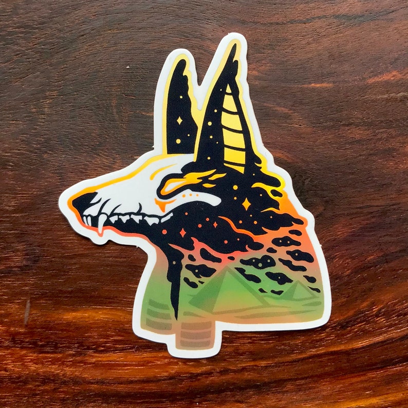 Pu&ograve; includere: Un adesivo nero e giallo con la testa di un jackal Anubis con un cielo stellato e un paesaggio verde e giallo sullo sfondo.