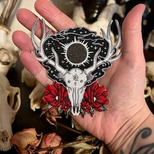 May include: An embroidered patch featuring a deer skull with antlers, a black sun, and red flowers. The patch is surrounded by a black background with white stars and clouds.