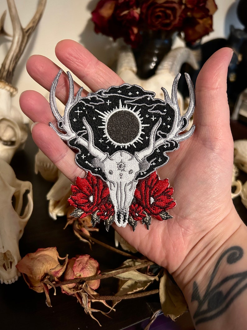 May include: An embroidered patch featuring a deer skull with antlers, a black sun, and red flowers. The patch is surrounded by a black background with white stars and clouds.