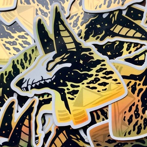 Pu&ograve; includere: Un mucchio di adesivi olografici neri e dorati con la testa di un chacal Anubis su uno sfondo di cielo stellato. Gli adesivi sono stampati con un gradiente di oro e giallo.