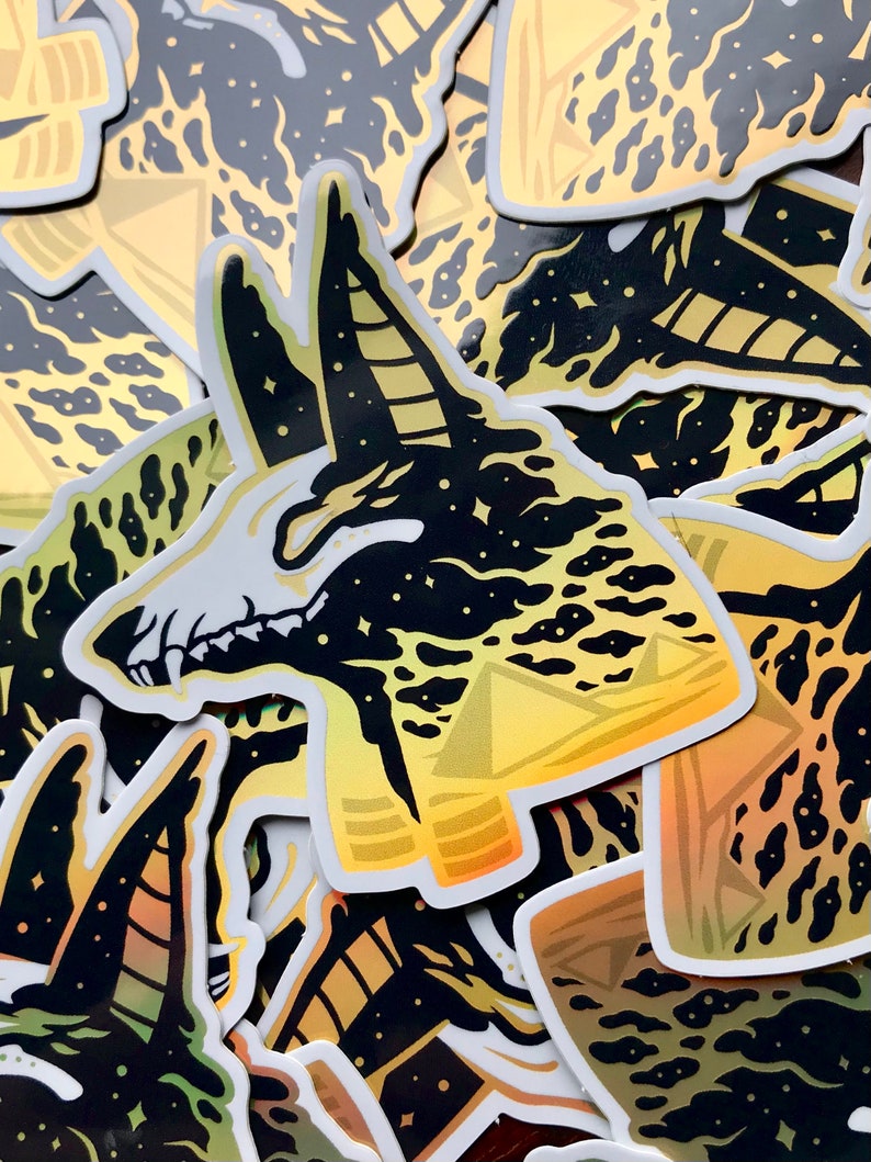 Pu&ograve; includere: Un mucchio di adesivi olografici neri e dorati con la testa di un chacal Anubis su uno sfondo di cielo stellato. Gli adesivi sono stampati con un gradiente di oro e giallo.