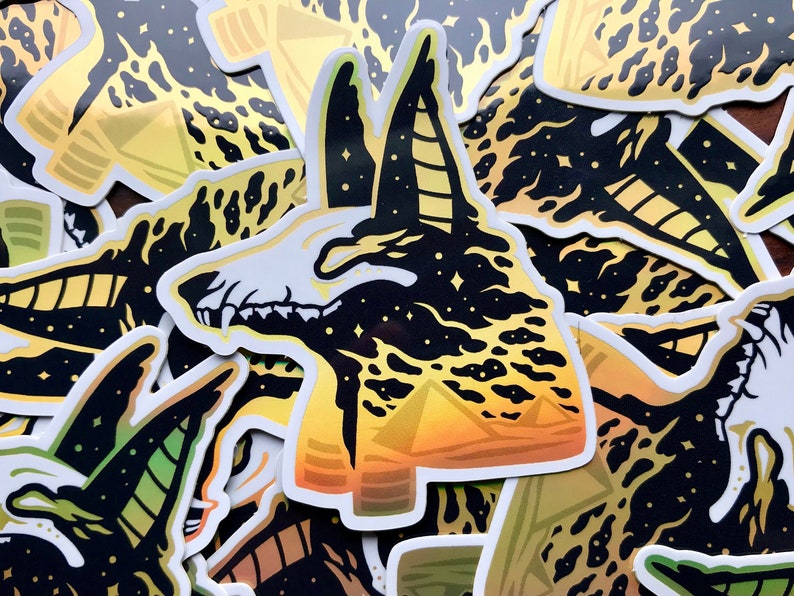 Pu&ograve; includere: Un adesivo nero e oro con un'immagine stilizzata di una testa di sciacallo Anubis su uno sfondo di cielo stellato e piramidi in primo piano.