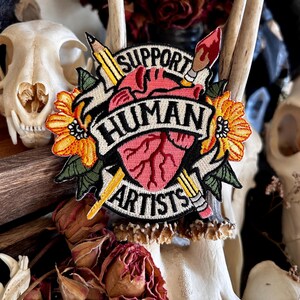 May include: A pink heart with a stylized anatomical design is surrounded by a yellow flower, a pencil, and a paintbrush. The heart is on a black banner with the text "Support Human Artists".