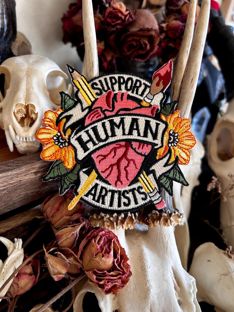May include: A pink heart with a stylized anatomical design is surrounded by a yellow flower, a pencil, and a paintbrush. The heart is on a black banner with the text "Support Human Artists".