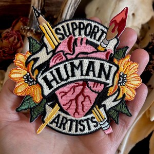 May include: Embroidered patch with a pink heart, a pencil, a paintbrush, and yellow flowers. The text reads "Support Human Artists".