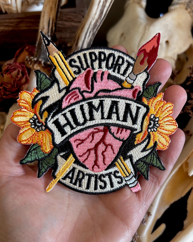 May include: Embroidered patch with a pink heart, a pencil, a paintbrush, and yellow flowers. The text reads "Support Human Artists".
