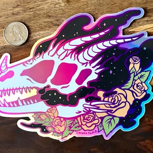May include: A holographic sticker featuring a pink and blue dragon skull with a horn, surrounded by roses and a starry night sky. The sticker is signed "Amanda Tillacz".