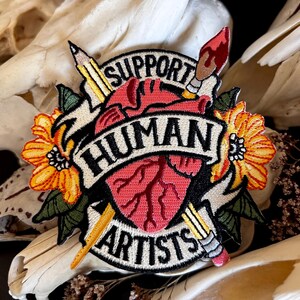May include: A black and white embroidered patch with a red heart, two pencils, a paintbrush, and yellow flowers. The text on the patch reads "Support Human Artists".