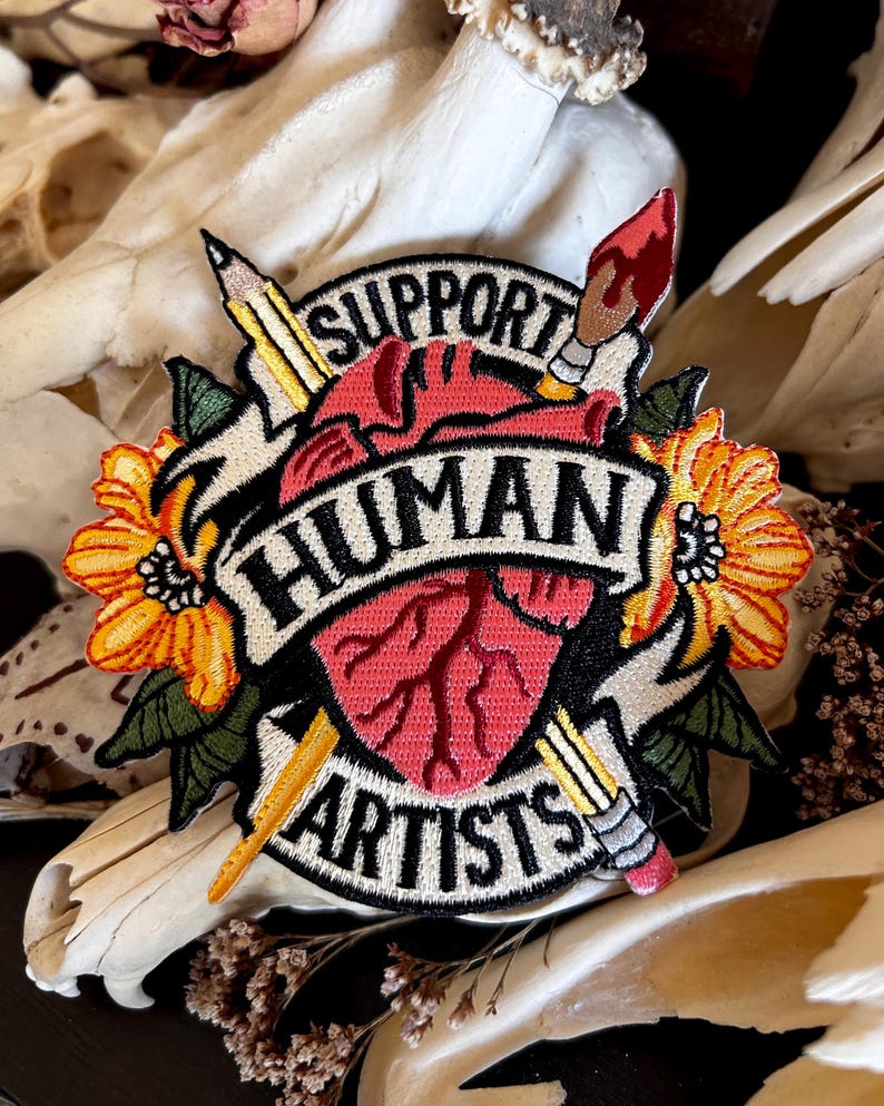 May include: A black and white embroidered patch with a red heart, two pencils, a paintbrush, and yellow flowers. The text on the patch reads "Support Human Artists".
