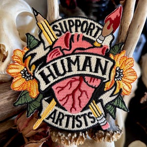 May include: Embroidered patch featuring a heart, two pencils, a paintbrush, and flowers. The heart has red veins and is surrounded by a banner that reads "Support Human Artists".