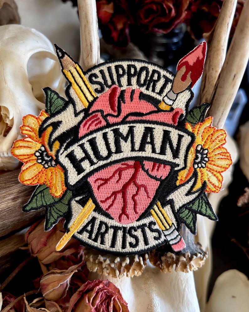 May include: Embroidered patch with a heart, two pencils, a paintbrush, and flowers. The heart has red veins and is surrounded by a banner that reads "Support Human Artists".