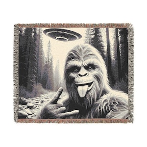 May include: Black and white woven throw blanket depicting a Sasquatch making a rock and roll hand gesture with a UFO above. The background shows a forest scene with tall trees and a rocky path.