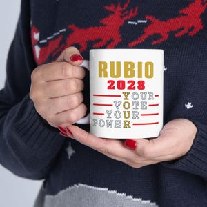 May include: White ceramic mug featuring the name "RUBIO" in gold lettering above the year "2028" in red. Below, the words "YOUR - VOTE - YOUR - POWER" are printed in red and gold. The mug is held by a person wearing a dark blue sweater.