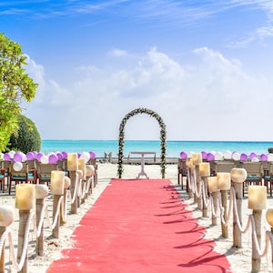 Peut inclure: Une c&eacute;r&eacute;monie de mariage sur la plage avec des chaises blanches, une arche blanche d&eacute;cor&eacute;e de fleurs violettes et un tapis rouge menant &agrave; l'arche.