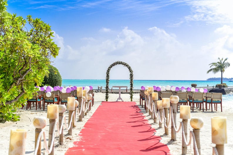 Peut inclure: Une c&eacute;r&eacute;monie de mariage sur la plage avec des chaises blanches, une arche blanche d&eacute;cor&eacute;e de fleurs violettes et un tapis rouge menant &agrave; l'arche.