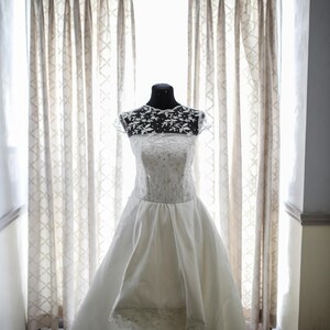Peut inclure: Une robe de mari&eacute;e blanche avec un corsage en dentelle et une jupe ample. La robe est expos&eacute;e sur un mannequin devant une fen&ecirc;tre avec des rideaux transparents.