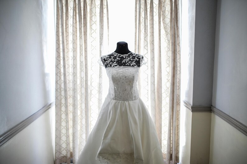 Peut inclure: Une robe de mari&eacute;e blanche avec un corsage en dentelle et une jupe ample. La robe est expos&eacute;e sur un mannequin devant une fen&ecirc;tre avec des rideaux transparents.