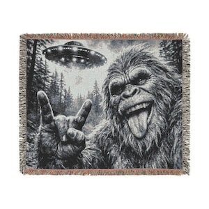 May include: A woven throw blanket with a black and white image of a Bigfoot character giving the rock and roll hand sign, with a UFO above. The blanket has a fringed edge and a forest scene.