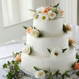 Peut inclure: Un g&acirc;teau de mariage blanc &agrave; trois &eacute;tages d&eacute;cor&eacute; de fleurs blanches et p&ecirc;che. Le g&acirc;teau est sur une nappe blanche et est entour&eacute; de verdure.