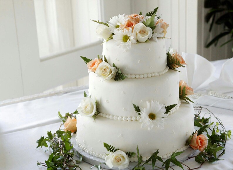 Peut inclure: Un g&acirc;teau de mariage blanc &agrave; trois &eacute;tages d&eacute;cor&eacute; de fleurs blanches et p&ecirc;che. Le g&acirc;teau est sur une nappe blanche et est entour&eacute; de verdure.
