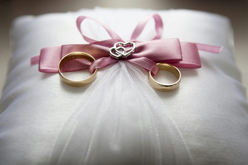 Peut inclure: Deux alliances en or sur un coussin blanc avec un ruban de satin rose et un charme en forme de c&oelig;ur argent&eacute;.