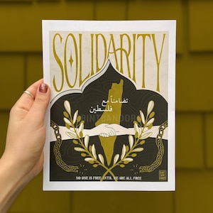 May include: A poster with the word "Solidarity" in large, stylized gold letters. The poster features a black silhouette of the country of Palestine with olive branches and a chain breaking apart. The text "No one is free until we are all free" is printed at the bottom of the poster.