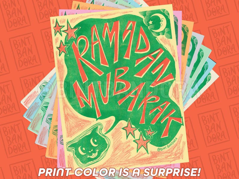 May include: A colorful greeting card with the text "Ramadan Mubarak" in red and green lettering. The card features a green cat with yellow eyes and a crescent moon in the top right corner. The card is surrounded by a stack of other cards with the text "Bint Ban Dora" repeated on each card.