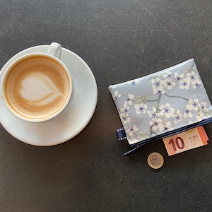 May include: A white ceramic cup with a latte, a blue and white floral zippered pouch with a 10 euro bill and a euro coin on a black surface.
