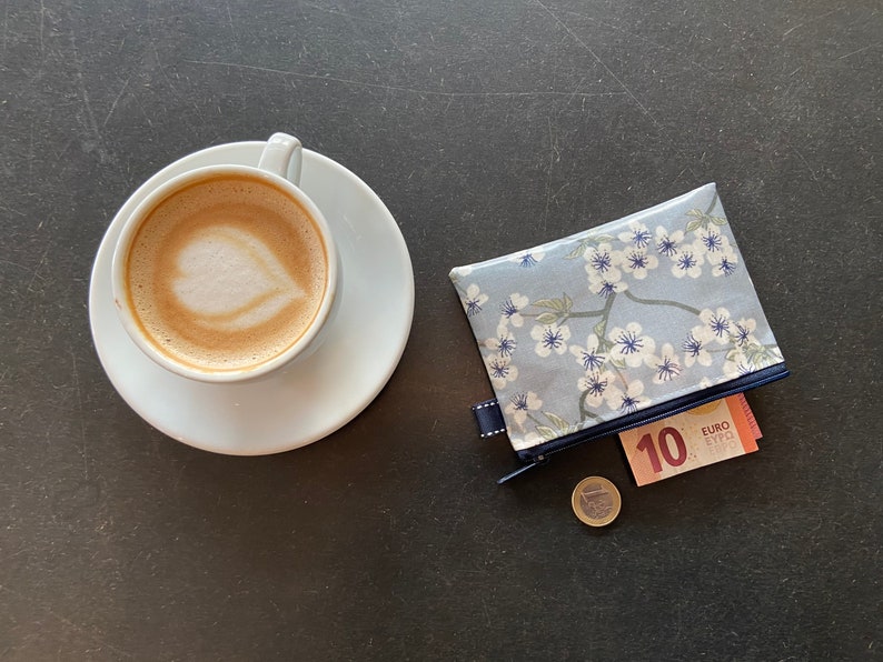 May include: A white ceramic cup with a latte, a blue and white floral zippered pouch with a 10 euro bill and a euro coin on a black surface.
